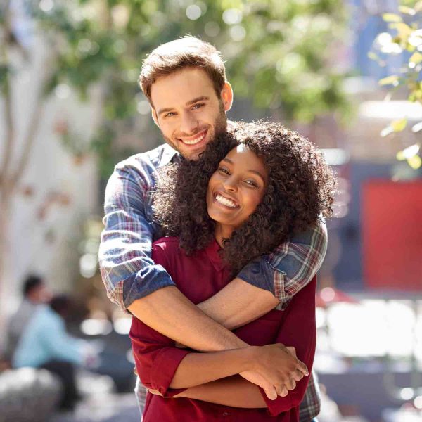Portrait of a smiling young man affectionately hugging his girlfriend from behind while standing together on a city sidewalk on a sunny day